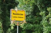 Ortsschild Neuenkirchen im Heidekreis vor grünem Wald im Hintergrund. Um das Ortsschild ist eine Kette aus Kartoffeln, Äpfeln und Mais gehängt.