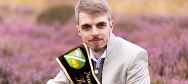 Kartoffelkönig 2026 Jannik Kartoffelkönig 2026 Jannik kniet in Heidelandschaft. Er trägt Anzug und eine Schärpe.