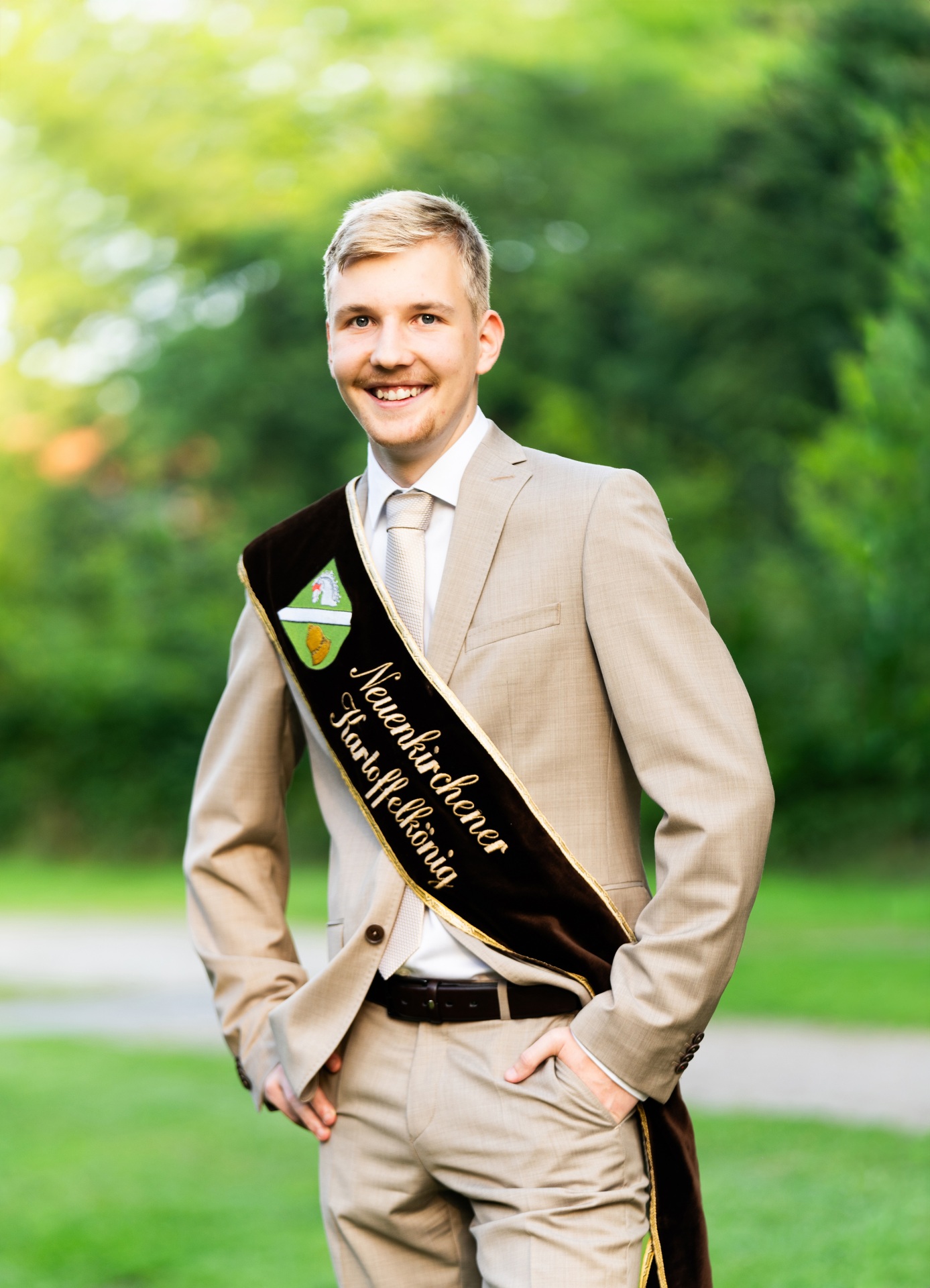 Portraitfoto des Kartoffelkönigs Georg Röhrs. Ein junger, blonder lächelnder Mann in beigem Anzug und einer braunen Scherpe mit der Aufschrift "Neuenkirchener Kartoffelkönig"