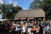 Blick auf die reetgedeckte Wagenremise auf dem Kartoffelfest 2025. Davor Tische und Bänke mit vielen Menschen. Getränkewagen hinten links im Bild.