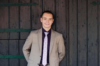 Proffesionelles Bild von Kartoffelkönig Tjarc-Ole. Beiger Anzug, lila Hemd und helle Krawatte. Lässig gegen ein dunkles Scheunentor gelehnt.